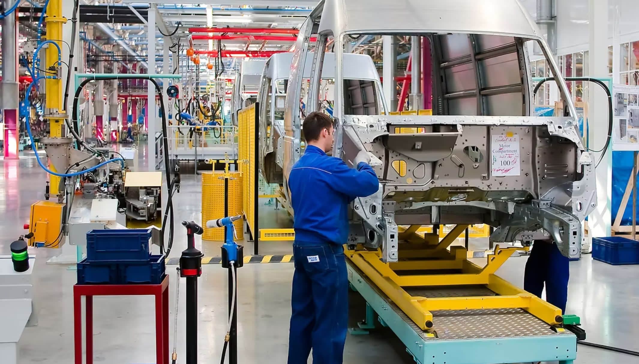A worker in blue overalls at a van body shell on an automotive assembly line.