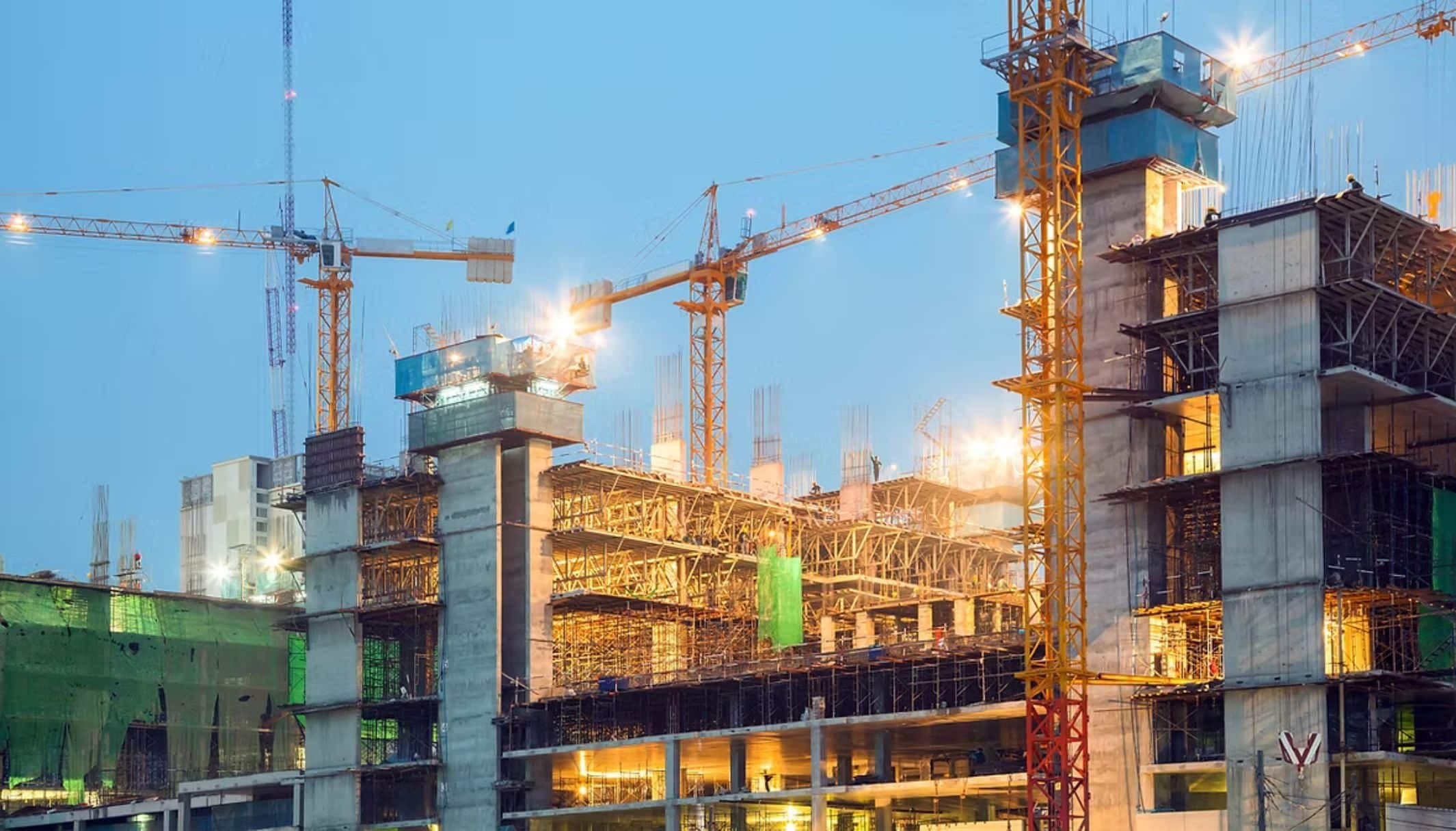 Tower cranes over a large concrete construction site, lit at dusk.