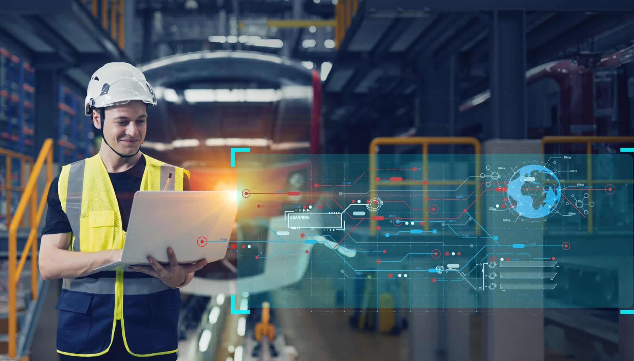 A worker in a hard hat and hi-vis vest using a laptop, with a data overlay across the factory behind.