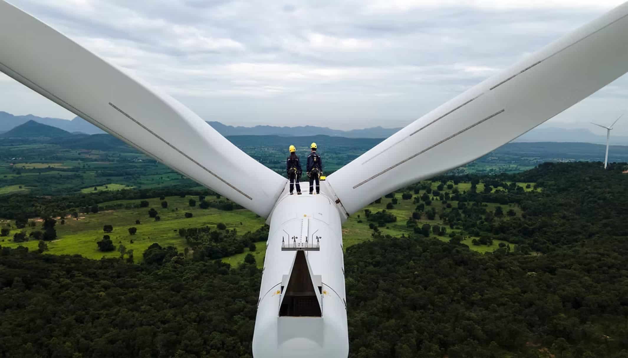 Two technicians standing on the nacelle of a wind turbine, with green hills below.